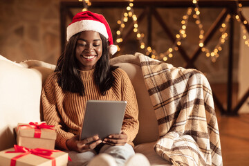 Online Christmas celebration. Black woman in Santa hat making video call, using tablet, shopping for Xmas presents