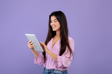 Happy armenian lady using digital tablet, looking at screen and smiling, standing over purple background, copy space