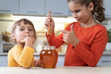 Happy children eating honey from a jar at home in the kitchen. funny girls lick honey from a spoon....