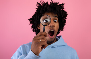 Shocked black teen guy looking through magnifying glass on pink studio background. Secret, doubt...