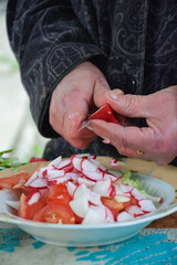 An old woman is cutting tomatoes and radishes into slices into a salad.