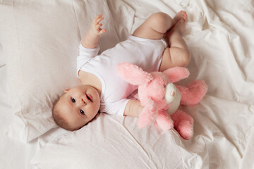 cheerful newborn baby in a white bodysuit lies on a white bed with a plush pink toy rabbit. products for children, toy. concept of a happy childhood and motherhood. child care. space for text.