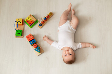 cheerful newborn baby in a white bodysuit lies on his back on the floor and plays with educational...