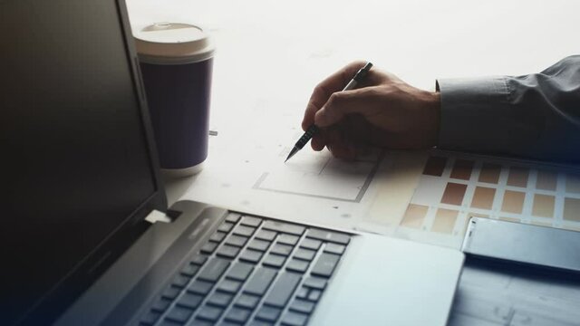 Composition from a laptop with a glass of coffee and a person's hand making a note on the drawing. Next to the table are samples of farb in the form of a palette shades.