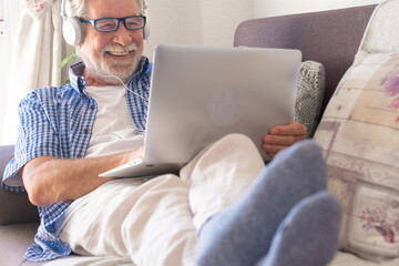 Attractive senior man at home sitting on sofa in video call with laptop computer. An elderly retiree enjoying free time and technological devices