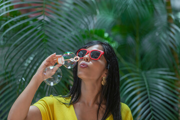 Woman in yellow dress and sunglasses with red frames blowing bubbles against a background of green plants outdoors