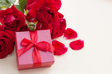Obraz na płótnie Canvas A red gift box for a ring with a red satin ribbon bow on a beige background next to a bouquet of red roses on green stems and red petals with a place for text