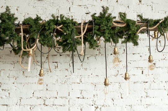 Festive Christmas hanging decoration with green spruce branches cords and light bulbs with golden cartridges on the background of a white brick wall