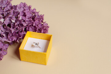 Yellow open box with a white gold ring with a white gemstone on a beige background with a purple lilac branch with a place for text