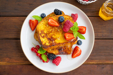 A stack of French toast on a plate with fresh berries, almond petals and honey on a dark wooden background. Delicious breakfast. Copy space.