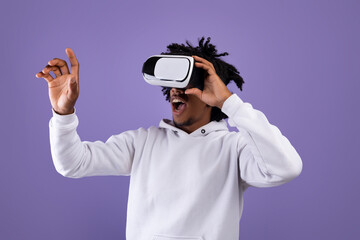 Amazed African American teen guy in VR headset using virtual reality, touching imaginary screen on violet background