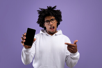 Irritated black teenager pointing at smartphone with empty black screen, offering mockup for app or website