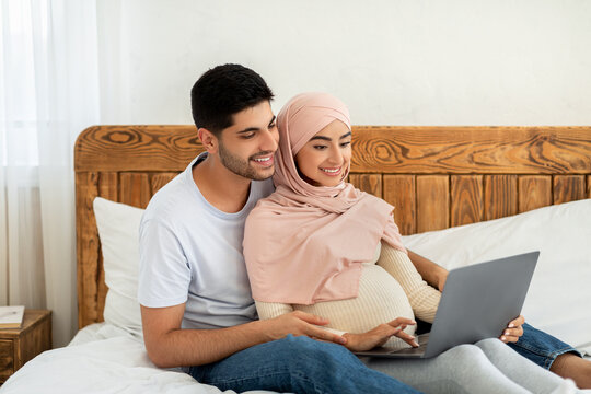 Cheerful pregnant arab couple using laptop computer, watching movie together while resting on bed - Powered by Adobe