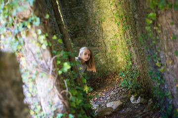 Cute little girl in old castle at forest. Curious girl with long fair hair peeking out from corner. Childhood, nature, fantasy concept