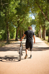 Back view of person with disability with bicycle. Curly man with mechanical leg walking in park on sunny day. Sport, disability concept