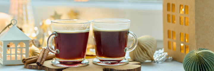 Hot espresso coffee with milk froth and cinnamon sticks on christmas decorated windowsill background banner. Light garland, interior winter composition, cold and cozy concept