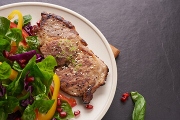 Grilled steak with fresh vegetable, sweet pepper, tomatoes, red onion, pink pepper and spices. Home made tasty food. Concept for tasty and healthy meal. Black stone background. Pork steak with salad