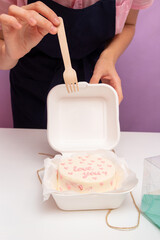 Bento cake with inscription "Love you" and hearts in plastic box-packaging. Above it, hand holds small wooden fork. Selective focus.