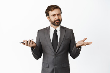 Whats wrong. Clueless businessman shrugging shoulders while holding cellphone, cant understand, dont know, standing over white background - obrazy, fototapety, plakaty