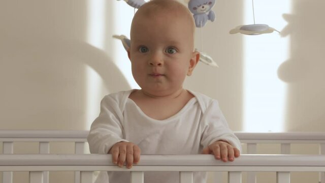 Cute smiling baby boy standing in cot at home, happy kid having fun learning to stand up in baby cot. High quality 4k footage