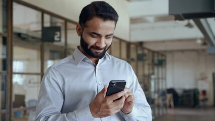 Happy indian ethnic businessman holding smartphone using mobile application program looking at cell phone working checking online digital data market corporate management tech standing in office. - Powered by Adobe