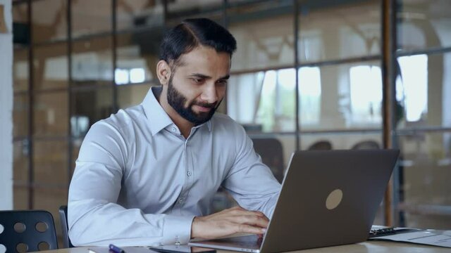 Indian ethnic business man employee professional manager using computer, typing, analyzing digital data working in modern office doing online data market analysis planning looking at laptop.