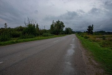 wet asphalt paved country road after rain with puddles. Stormy clouds ahead.