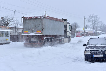 Winter. Snow falls. A large lorry is driving on a road that has not been cleared of snow.