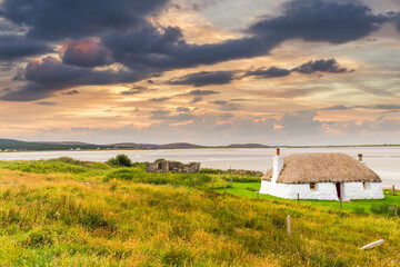 Traditionally built white cottage with thatched roof, next to the turquoise bay, with stormy cloudy dark skies above.island of North Uist, Scotland - obrazy, fototapety, plakaty
