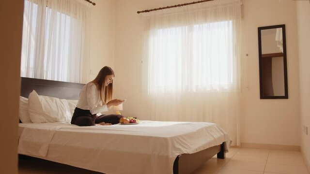 Happy young woman sitting on the bed with a plate of fresh fruits and taking pictures on a mobile phone. Woman traveler enjoying free time in a cozy hotel room. People and technology concept