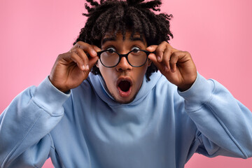 Shocked young black teen guy opening mouth in awe, touching eyeglasses, looking at camera on pink...