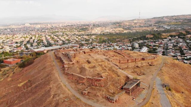 Aerial panning view triangle shape Erebuni fortress ruins on hilltop with Yerevan city panorama background . Old urartian fortified city . Important historical site in Armenia