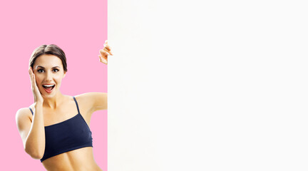 Photo of excited beautiful brunette woman in fitness wear showing blank banner paper signboard with copy space area. Fit girl with happy wide open mouth, peek out mock up white placard, over rose pink