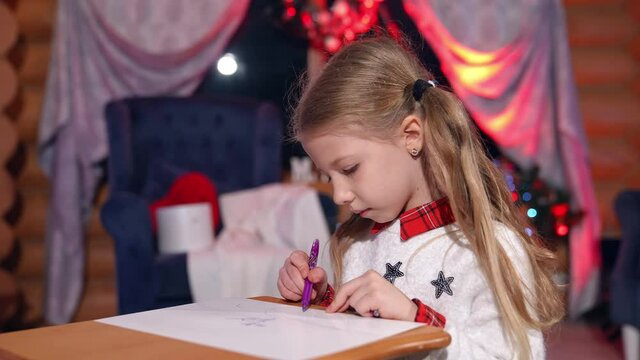 Little girl painting on Christmas tree background. Cute child drawing a picture to Santa before Christmas. New Year tree with bright lights in the room.