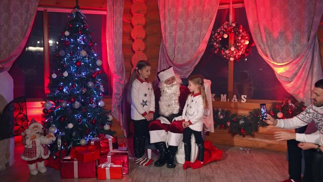 Santa Claus and little girls near Christmas tree. Children telling rhymes to Santa to get presents from him. Happy parents taking photos of children at Christmas.