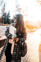 Beautiful young girl student with coffee, books and a backpack spending good time at the park, music with headphones