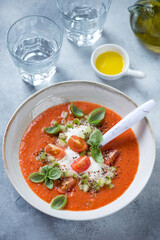 Tomato gazpacho soup with addition of burrata cheese and fresh green basil, vertical shot on a light-blue stone background