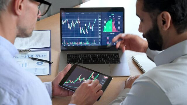 Two diverse traders brokers stock exchange market investors discussing crypto trading charts growth using digital tablet and laptop analyzing financial risks, investment profit, global rates forecast.