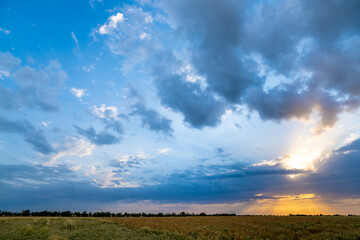 Obraz premium A picturesque sunset over an agricultural field. Blue sky and the sun behind beautiful clouds at sunset. The sun's rays break through the clouds.