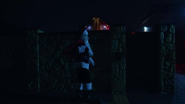 Santa Claus with red bag at night. Santa walks into front yard of house decorated with lights and garlands. Santa carrying gifts for children.