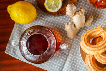 A teapot of strong infused tea on a napkin next to lemon, ginger and jam and donuts with cream top view.