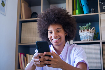 Fototapeta na wymiar Smiling african american woman with curly hairstyle sitting at office and text messaging using mobile phone. Happy businesswoman using mobile phone at workplace. 
