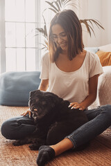 Portrait of beautiful woman in eyeglasses having fun with her pet pug dog sitting on floor in the living room of her house. Cheerful woman spending leisure time with her cute pet dog at home.