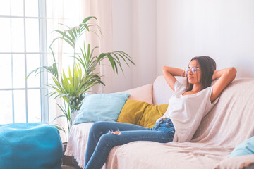 Happy young woman in eyeglasses relaxing on sofa with hands behind head in the living room of her house. Carefree and satisfied lady with torn jeans relaxing on couch at modern apartment.