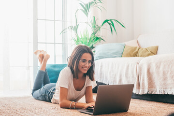 Happy young woman in eyeglasses typing using laptop while lying down on carpet in the living room of her house. Beautiful lady spending leisure time using laptop. Freelancer working from home.