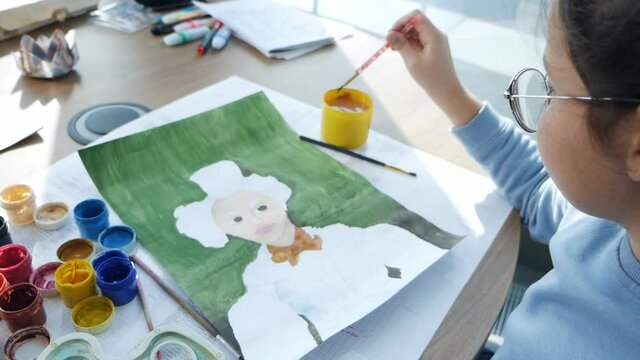 Creative child learns to draw on paper. A little girl sitting at the table draws with a brush and colored liquid paints from jars.Slow motion
