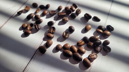 Coffee beans, scattered around on white wood background on sunlight under windows shades shadows,...