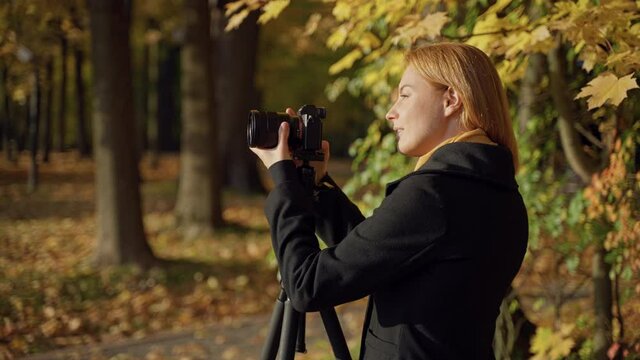 Caucasian young red-haired woman photographer In a black coat takes photos of landscapes in an autumn park with a mirrorless camera with a wide-angle lens on tripod, looking through viewfinder, smiles