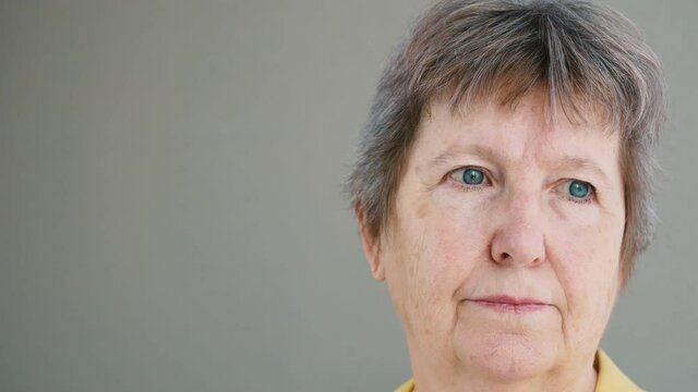 Close-up face of an older adult woman, with blue eyes. The hair is gray and the face is marrowed and the skin is covered with age spots. Calm face of grandma