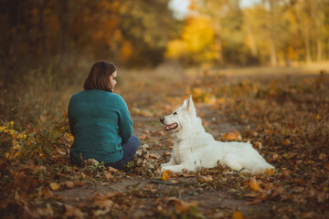woman with a dog white swiss shepherd sitting on a path near forest in autumn